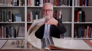 video still of Stephen Bury seated, flipping large scrapbook in library setting