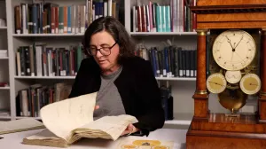 video still of Sally Brazil looking at book next to a table clock