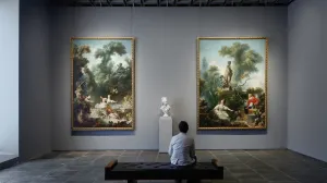 Man seated in a gallery with two large, colorful Rococo paintings and a marble bust