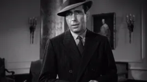 Man in a fedora stands in front of furniture and a wall with sconces and a framed portrait