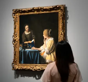 Woman looking at Vermeer’s “Mistress and Maid” in a gallery with gray walls.