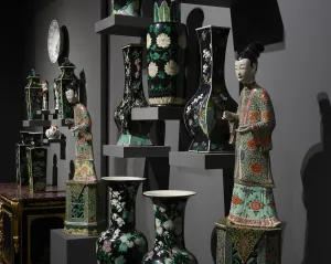 Installation view detail of a porcelain gallery with many black, green, and white porcelain objects on shelves, with two ornately decorated porcelain figures of women on stands featured.