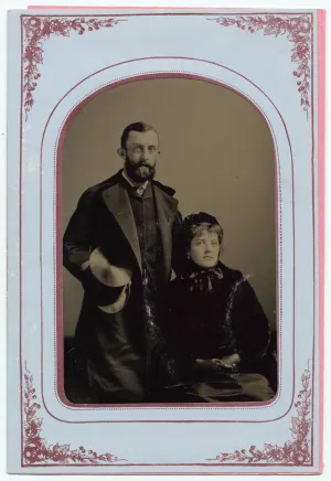 Black-and-white portrait photo in a decorative frame of a young couple, the bearded man standing in a coat and the woman seated, wearing a hat