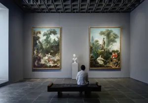 Man in a powder-blue coat sitting in a gallery with gray walls featuring two large, colorful Rococo paintings flanking a white marble bust of a woman