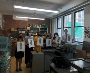 Six people in face masks posing for a photo holding large paper prints with the initials CBA