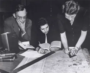 Two women and a man at a desk holding books and marking a map with pencils and rulers