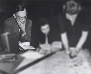 Three figures at a desk. A man at left is highlighted, holding a book with his hand to his chin.
