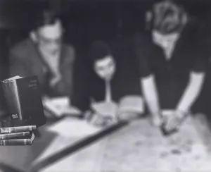 Three people at a desk. At left, a group of books on the table is highlighted.