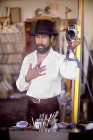 Man in a black hat holding a camera up to a mirror in an art studio
